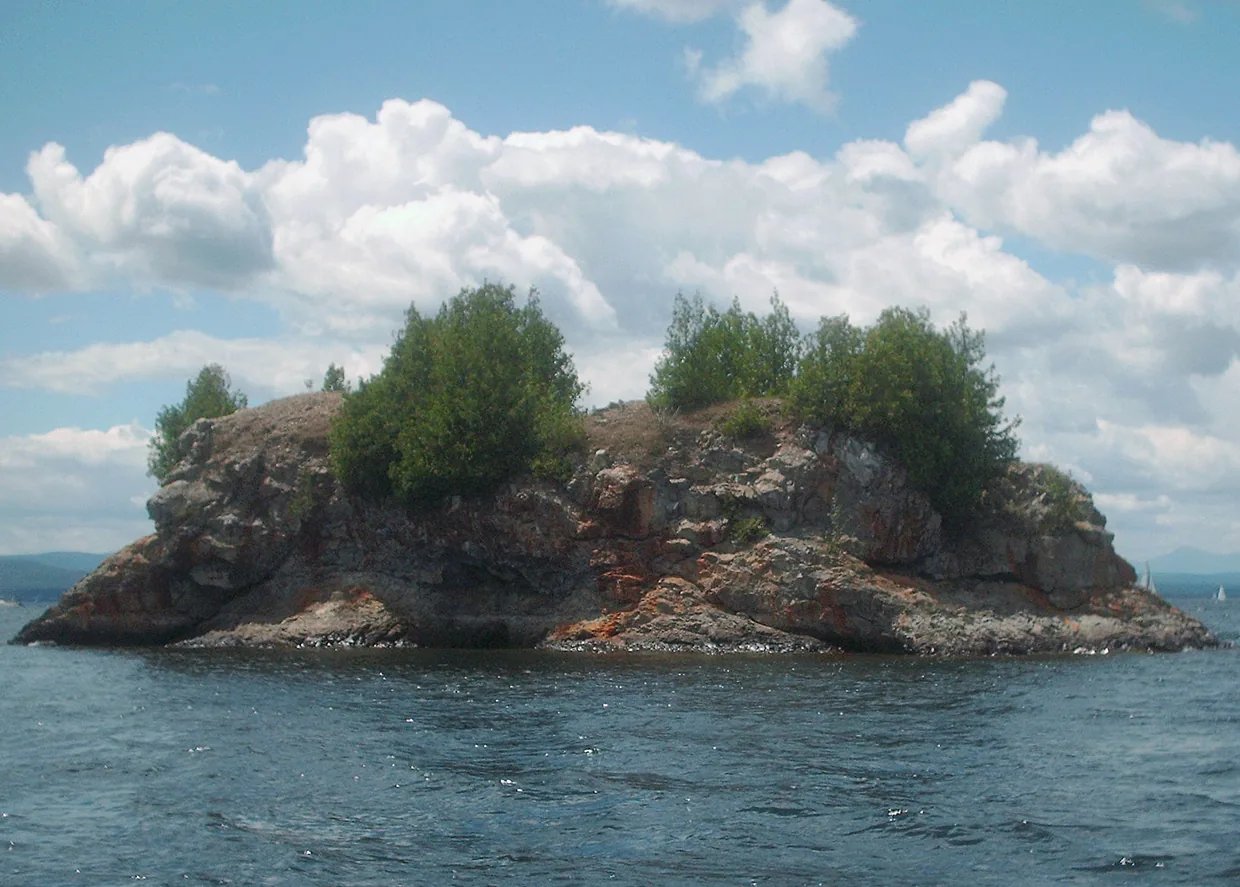 Carleton's Prize from the water: a steep-sided rock with a level top and a few trees on
      it, standing alone in open lake, with orange staining across the rock face.