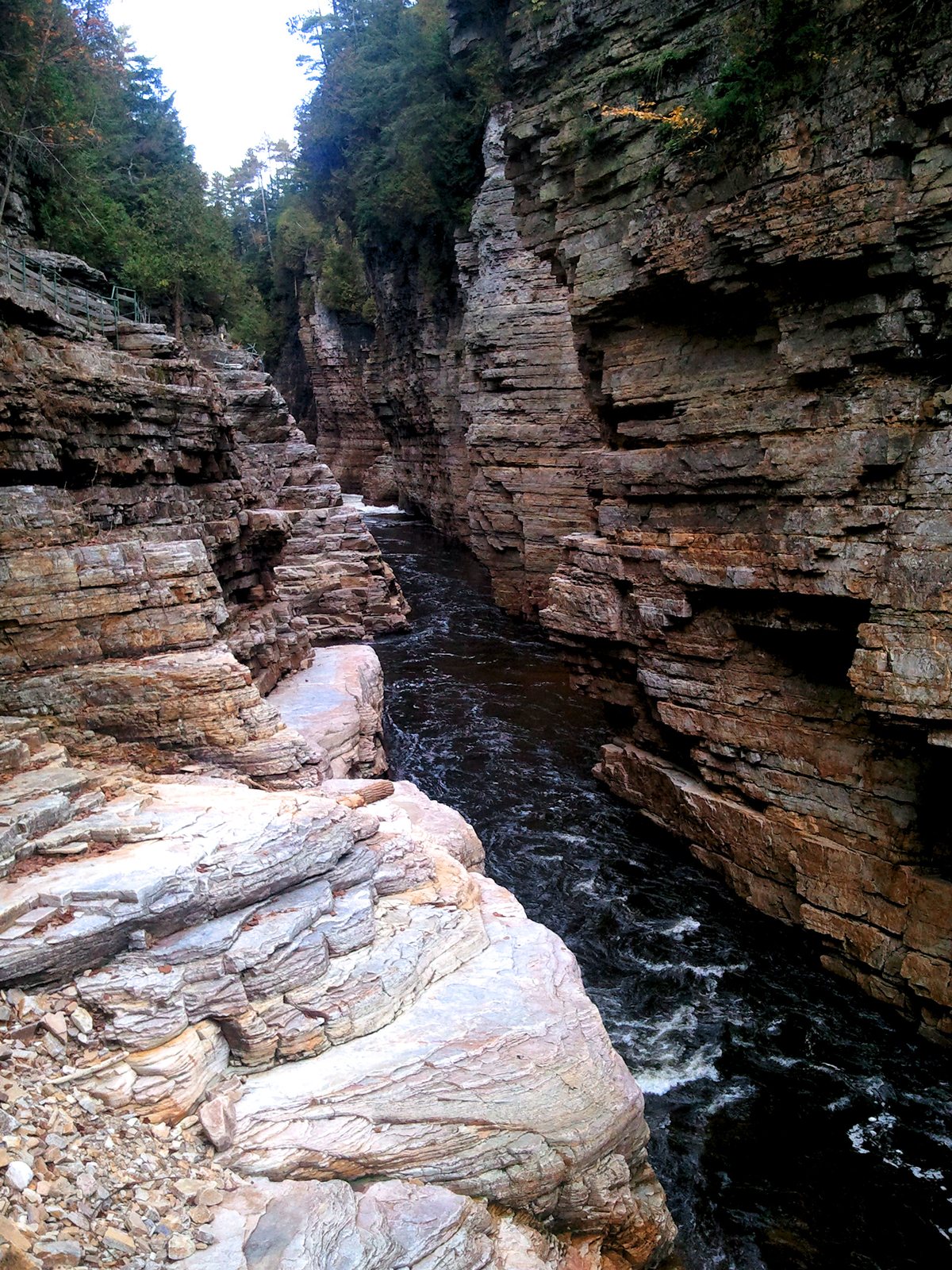 A narrow river gorge between vertical walls of layered orange-brown sandstone, seen from a path along the water