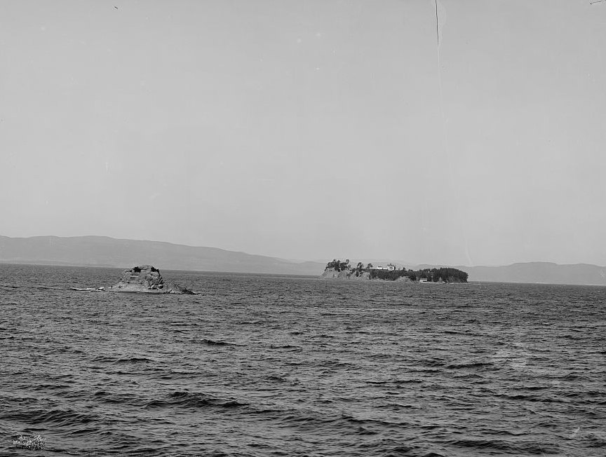 A black and white photograph across open water: a
    small bare rock breaking the surface at left, a low wooded island with buildings on it at right,
    and the ridge of the far shore behind them.